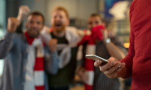 A man in the foreground looks at his phone during a sports match as his friends cheer in the background