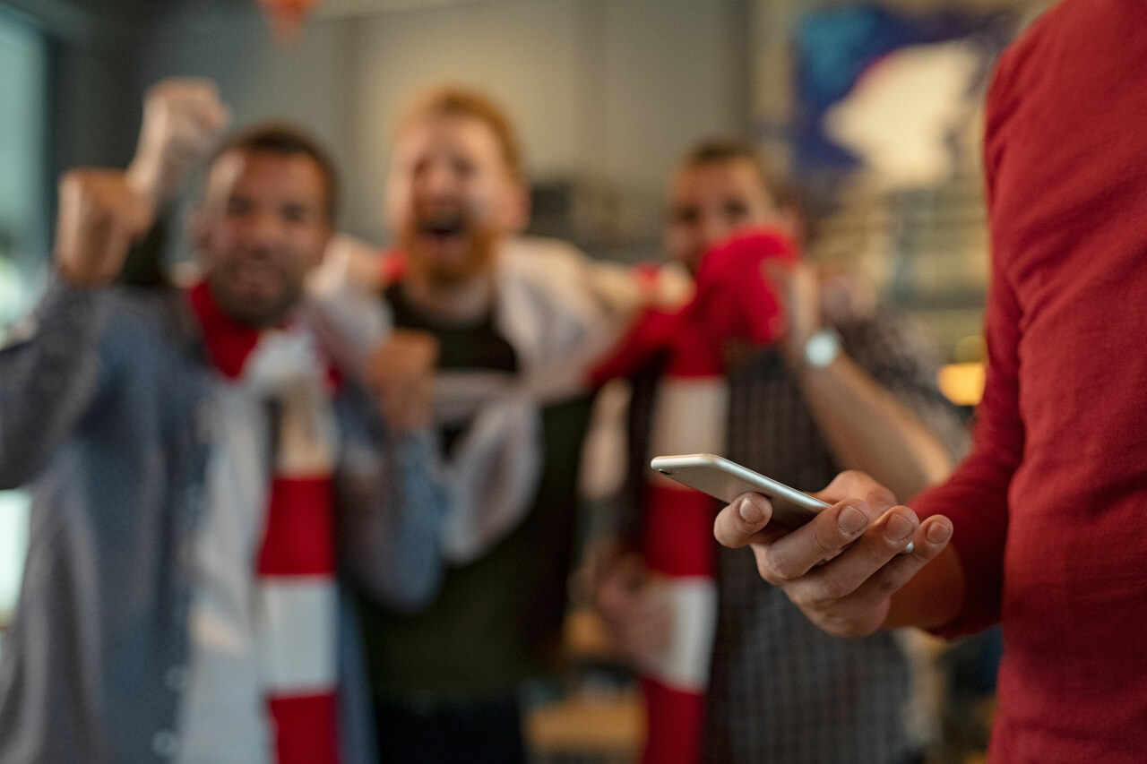 A man in the foreground looks at his phone during a sports match as his friends cheer in the background